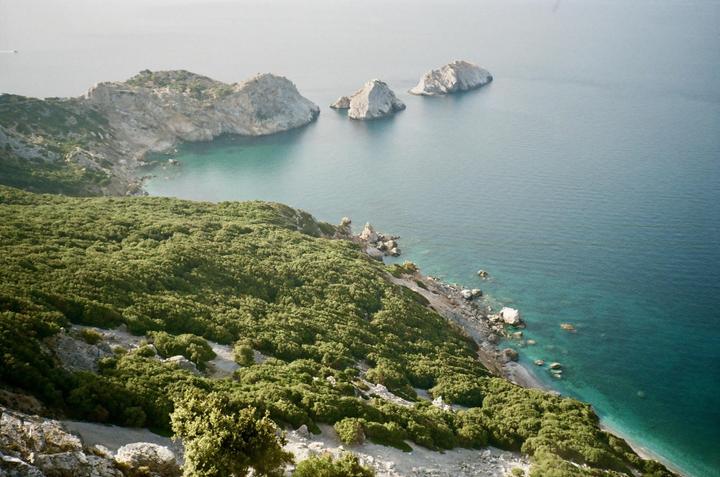 A high-angle landscape shows a lush green hillside descending to a rocky coastline and turquoise water with three small islands in the distance.