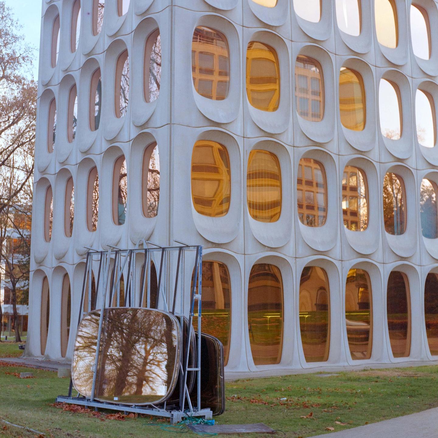 A building with many oval windows reflects trees and other buildings.