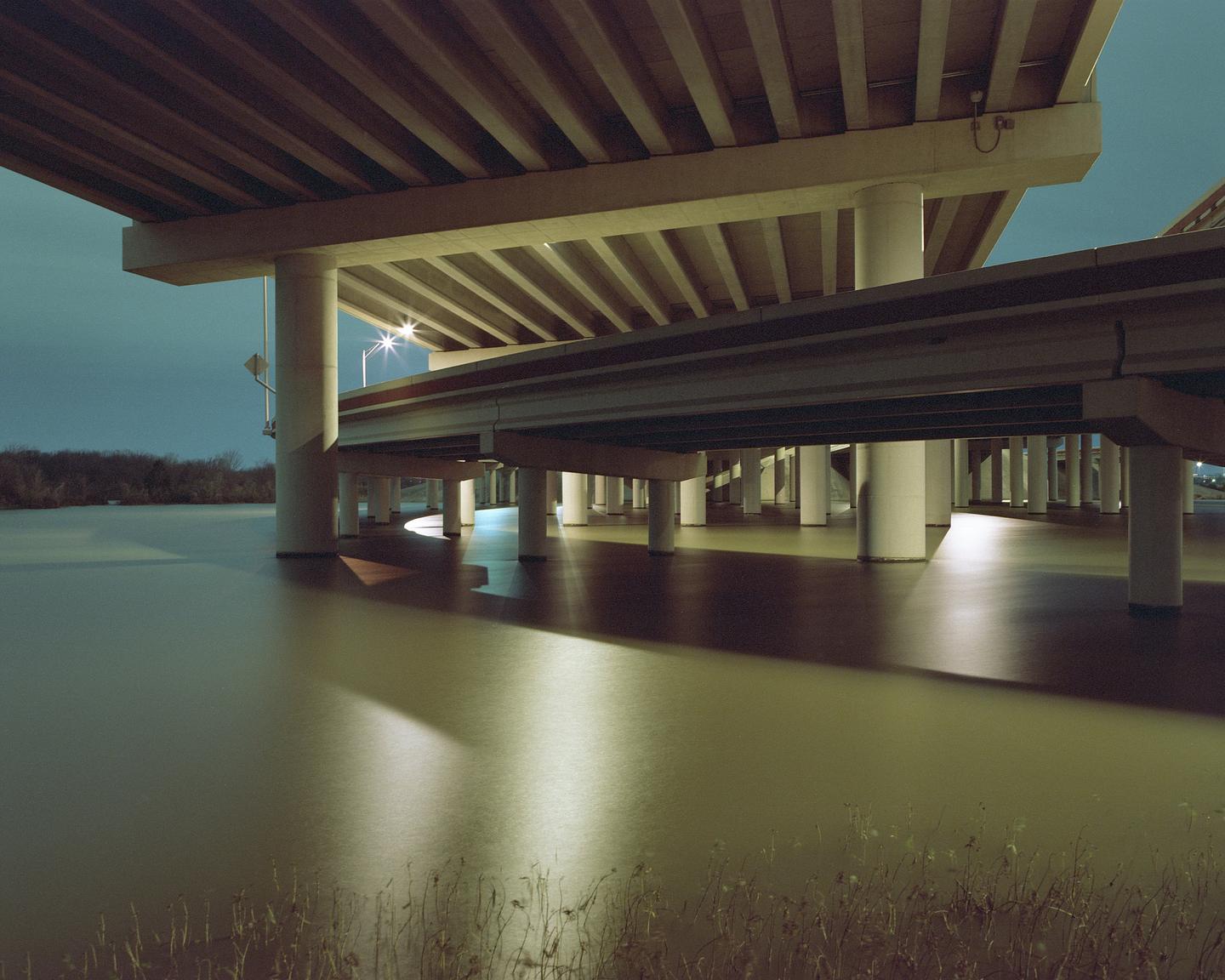 A highway overpass with many support pillars stands over a body of water at night.