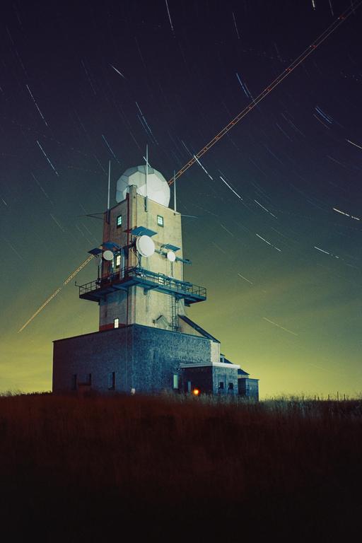 A tall building with a radar dome on top stands under a night sky filled with star trails and a light streak.