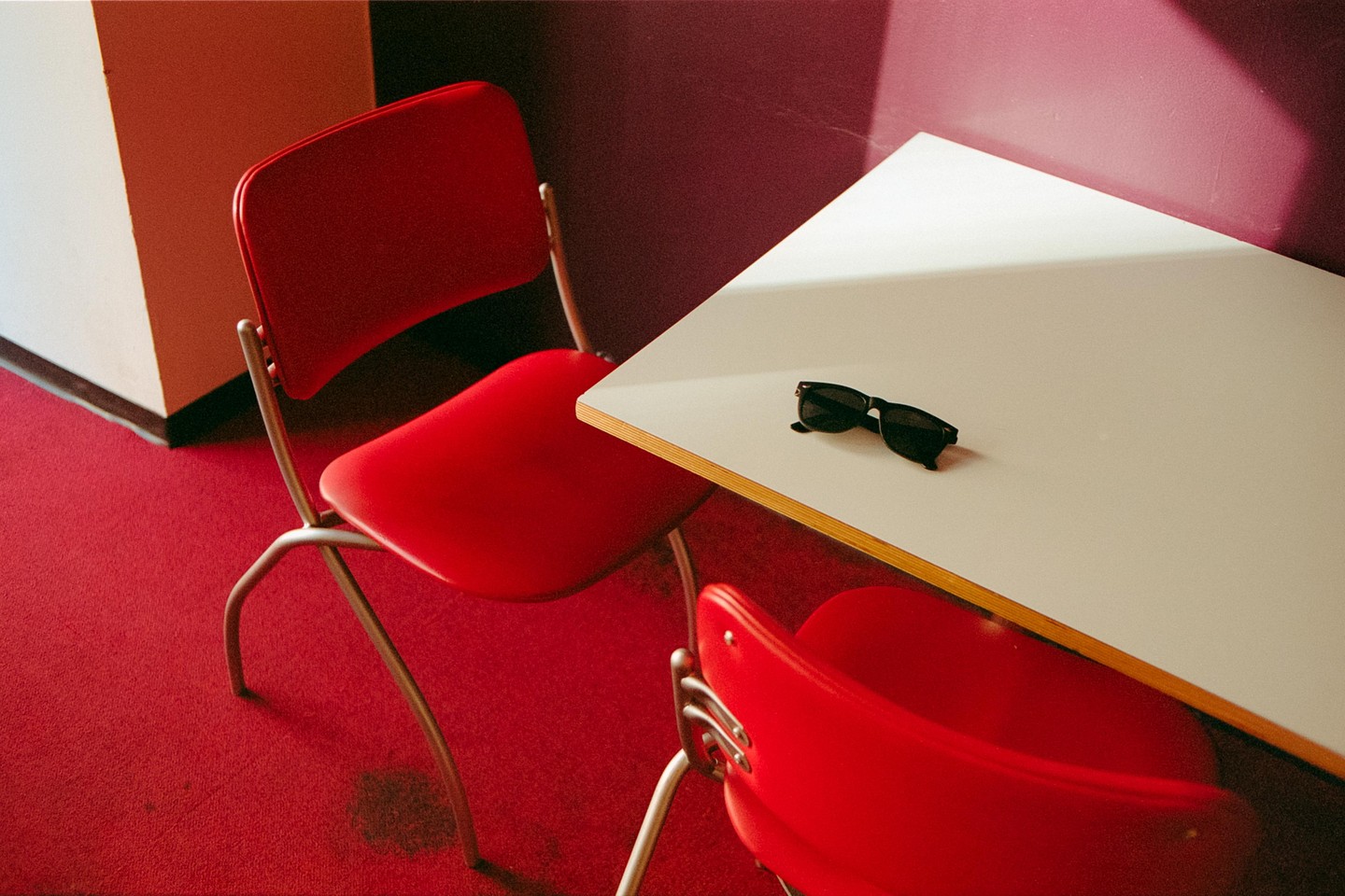A pair of sunglasses rests on a white table next to two red chairs on a red carpet.