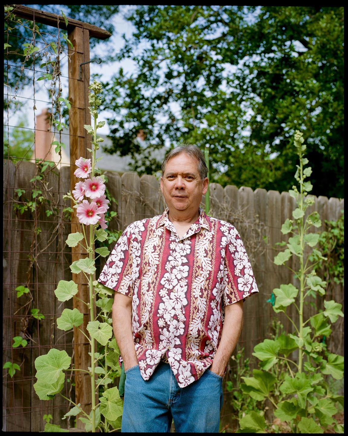 A man wearing a floral shirt and jeans stands in a garden next to a tall plant with pink flowers.
