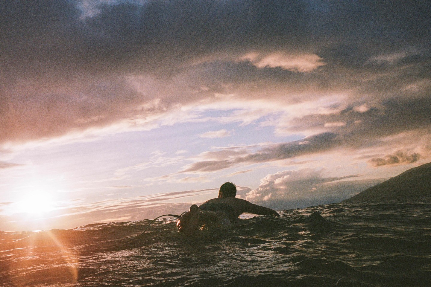 A surfer waits in the ocean at sunset with mountains in the background.