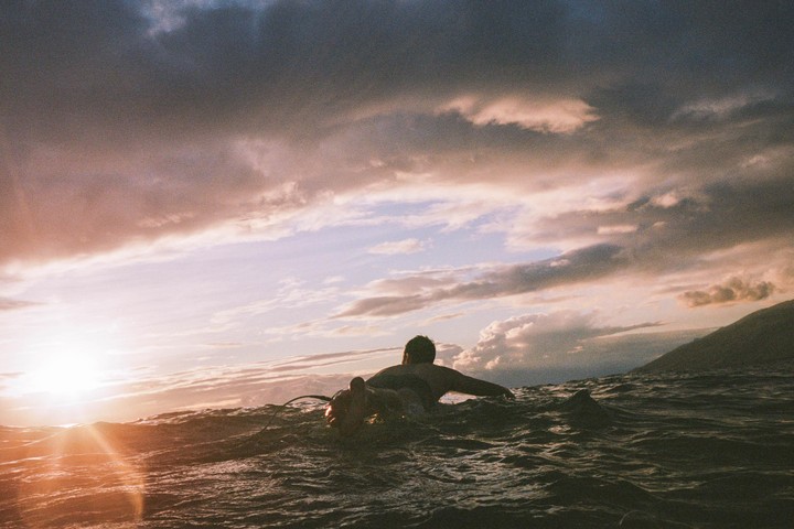 A surfer waits in the ocean at sunset with mountains in the background.