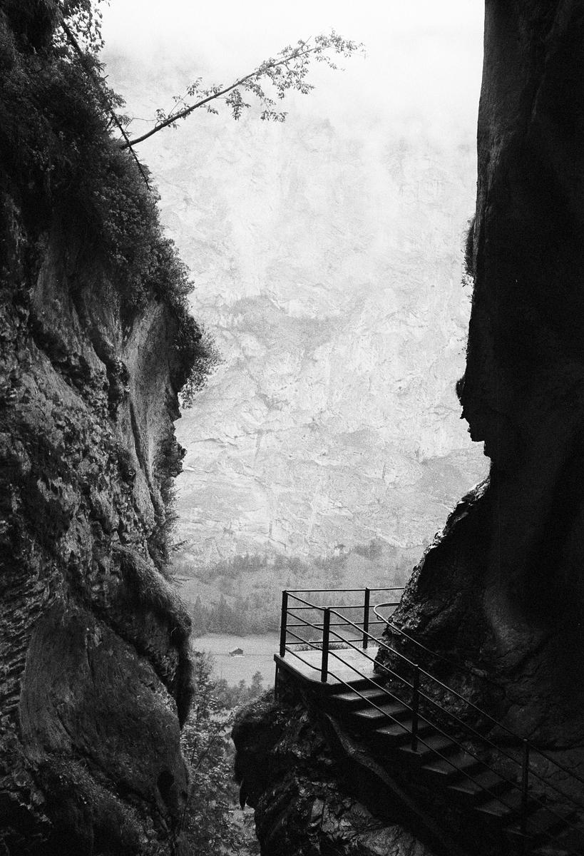 A stairway with a railing is built into the side of a cliff, overlooking a valley with a body of water and distant mountains.