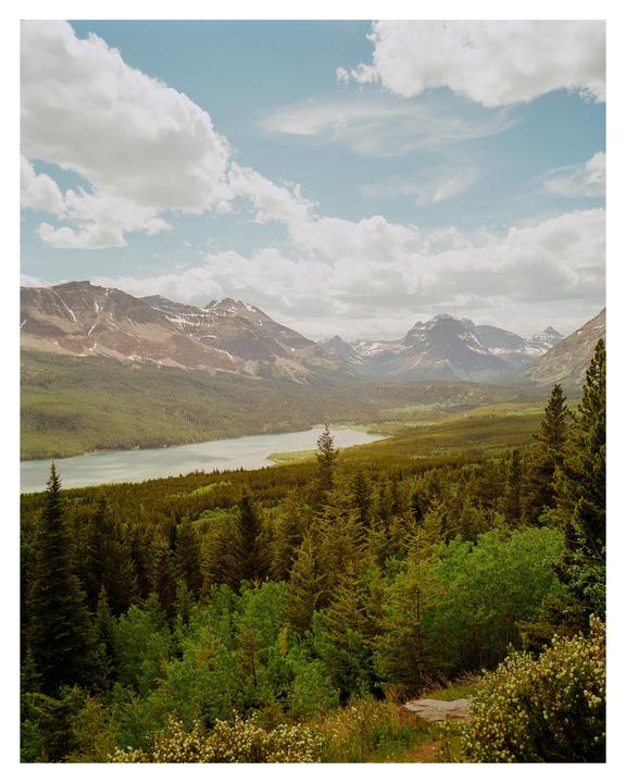 A wide landscape shows a lake surrounded by a dense forest with mountains in the background under a cloudy sky.
