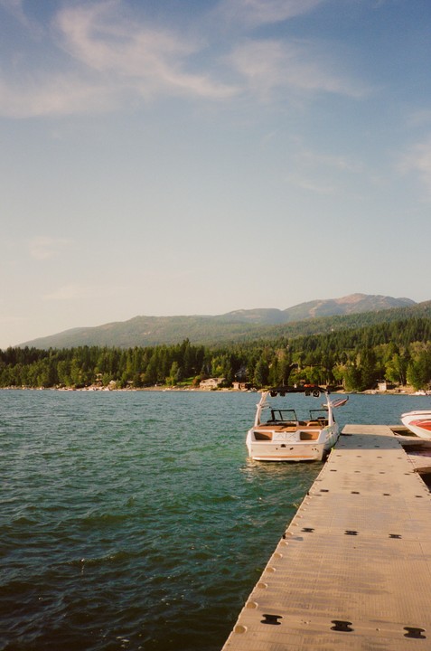 A boat is docked next to a floating pier on a lake surrounded by a forest and mountains under a clear sky.