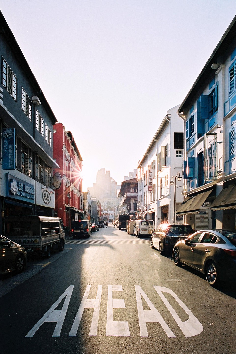 A street with parked cars and buildings on both sides has the word "AHEAD" painted on the asphalt, with the sun shining brightly at the end of the street.