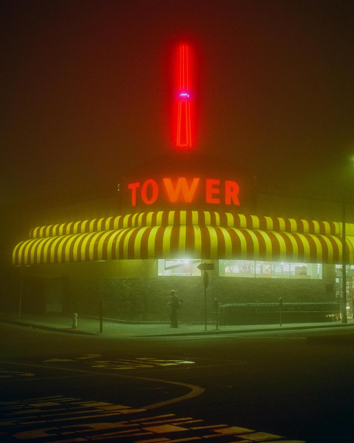 A person stands outside a store with a red and yellow striped awning and a neon sign that reads "Tower" in red.