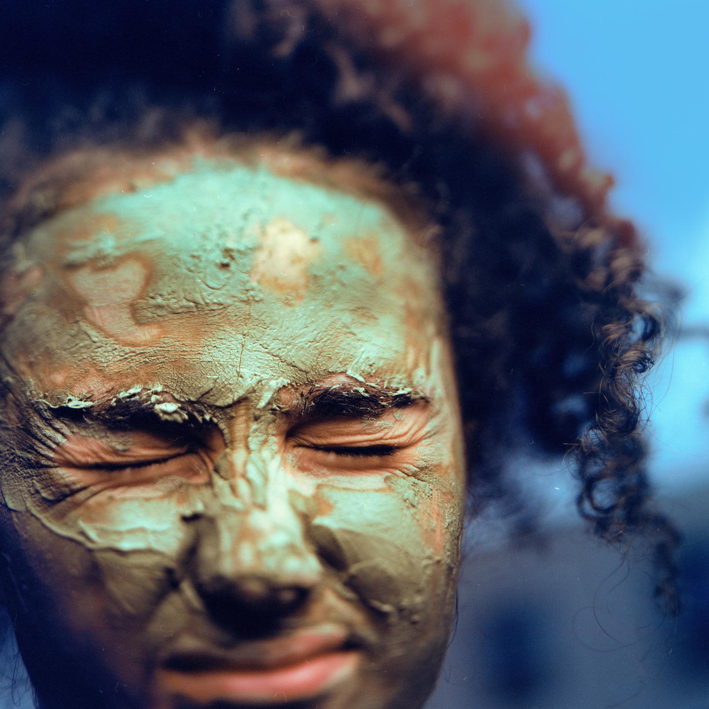 A woman with curly hair has a mud mask on her face and her eyes are closed.