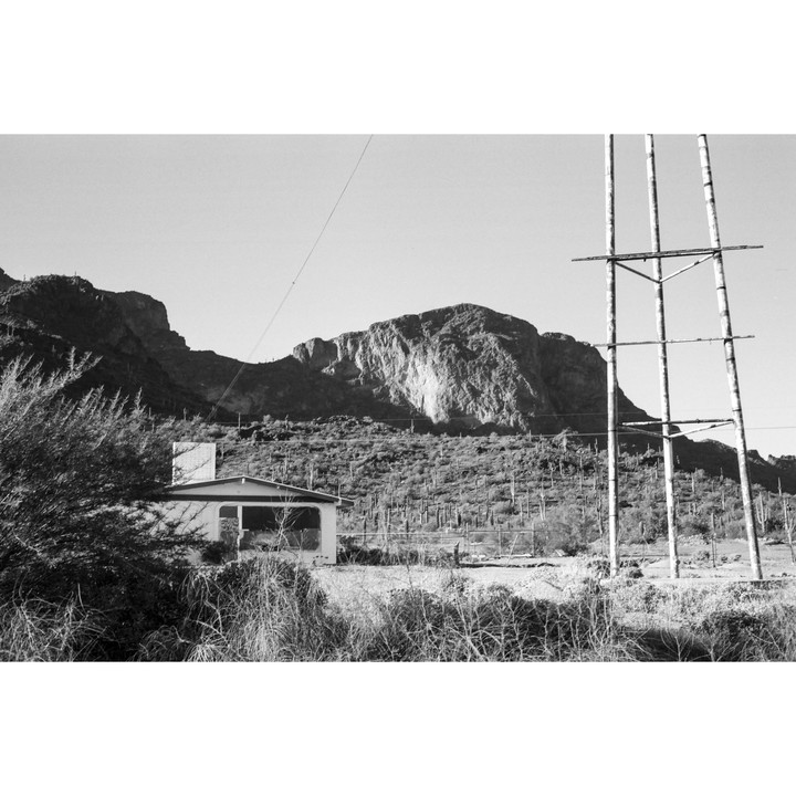 A small building sits in a desert landscape with mountains and cacti in the background.