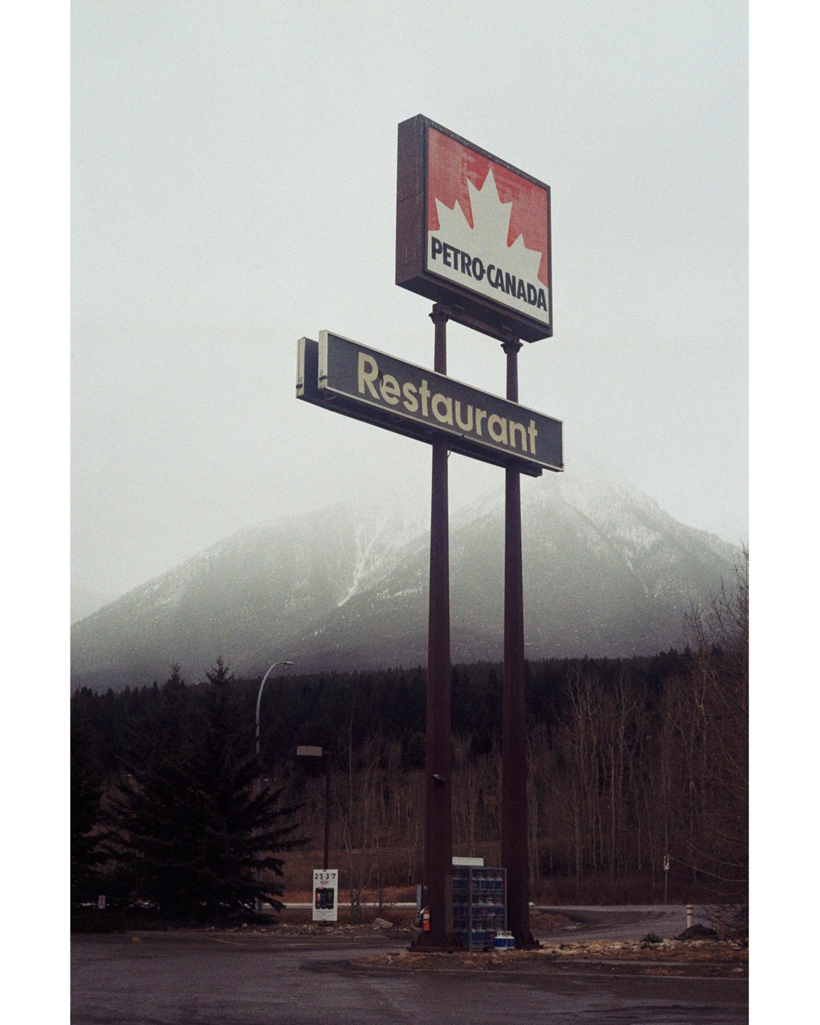 A Petro-Canada sign stands in front of a restaurant sign with a mountain and forest in the background.