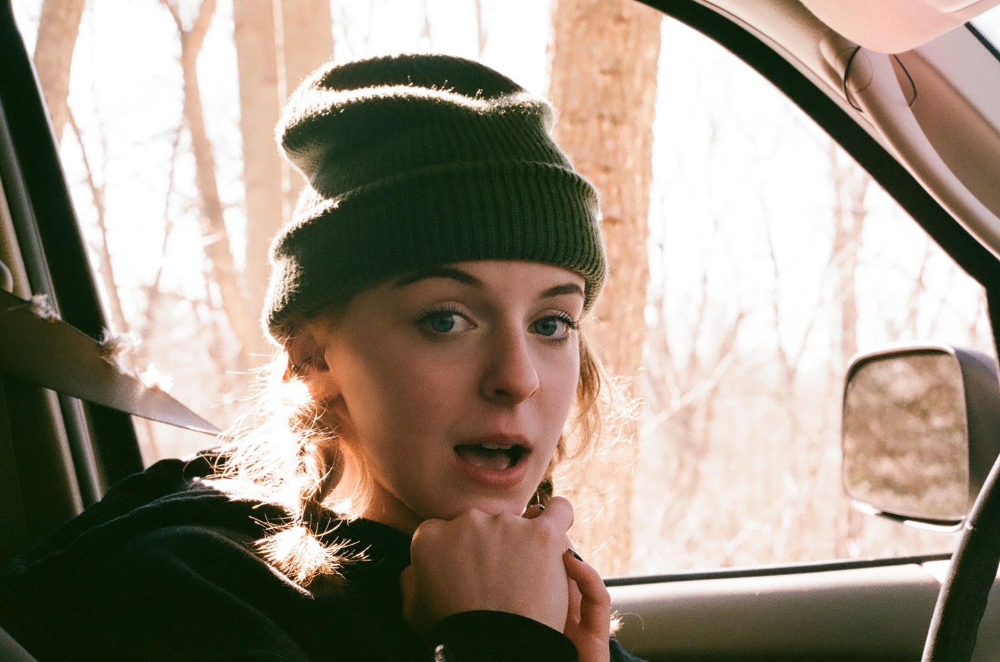 A young woman wearing a beanie looks surprised while sitting in a vehicle.