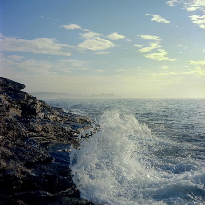 A wave crashes against a rocky shore under a partly cloudy sky.