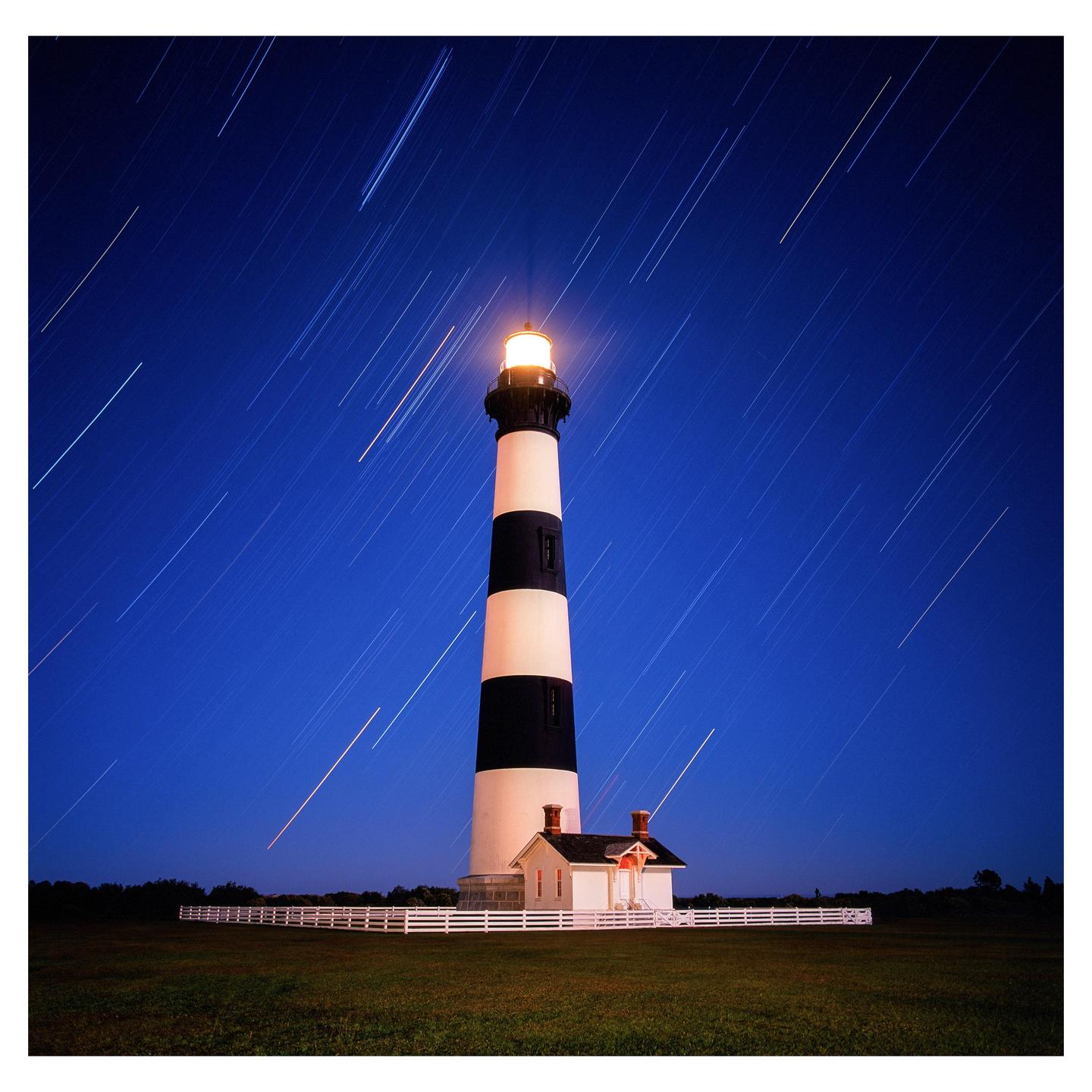 A black and white striped lighthouse stands tall against a deep blue night sky with star trails.