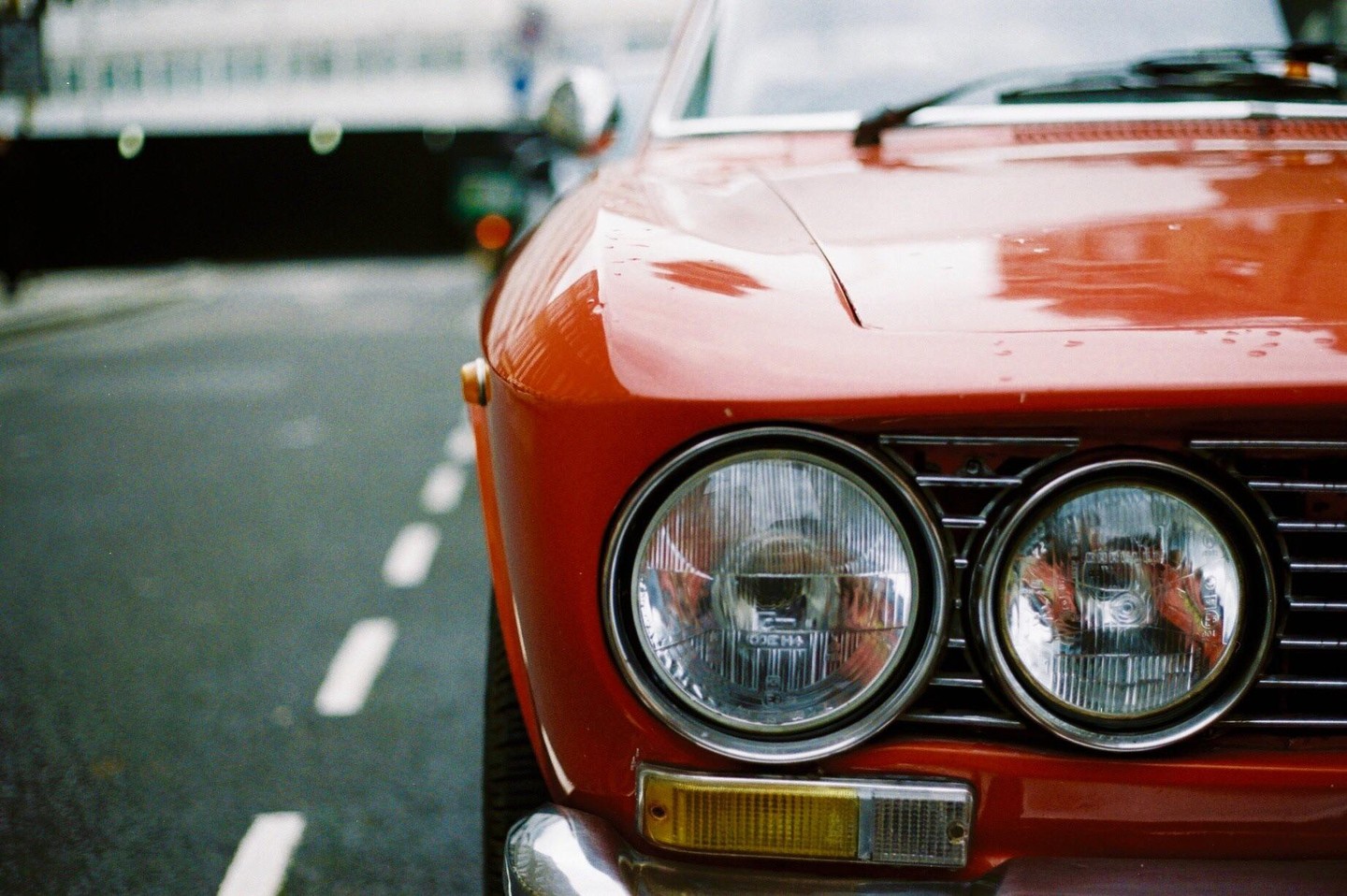 The front of a red vintage car with round headlights is parked on a wet street with white dashed lines.