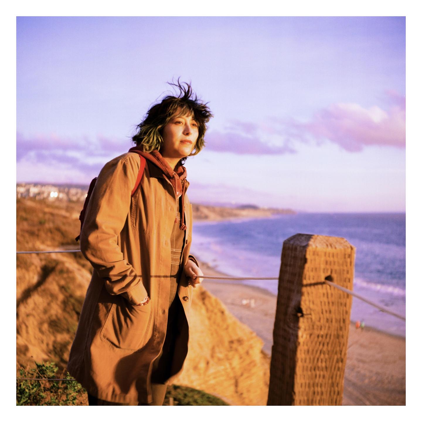 A woman stands on a cliff overlooking a beach and ocean at sunset.