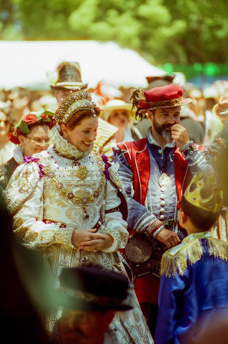 A woman in elaborate period costume and a man in a red hat and period costume look at a child in a blue costume and gold crown.