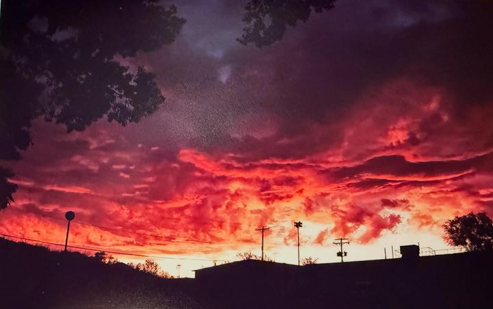 A vibrant red and orange sunset fills the sky above a silhouetted landscape with trees and utility poles.