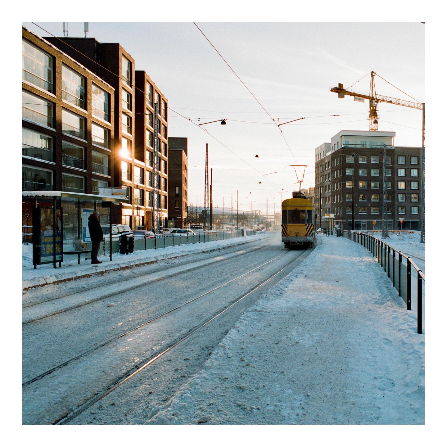 A man stands at a snowy tram stop with a tram approaching in the distance, surrounded by buildings and overhead wires.