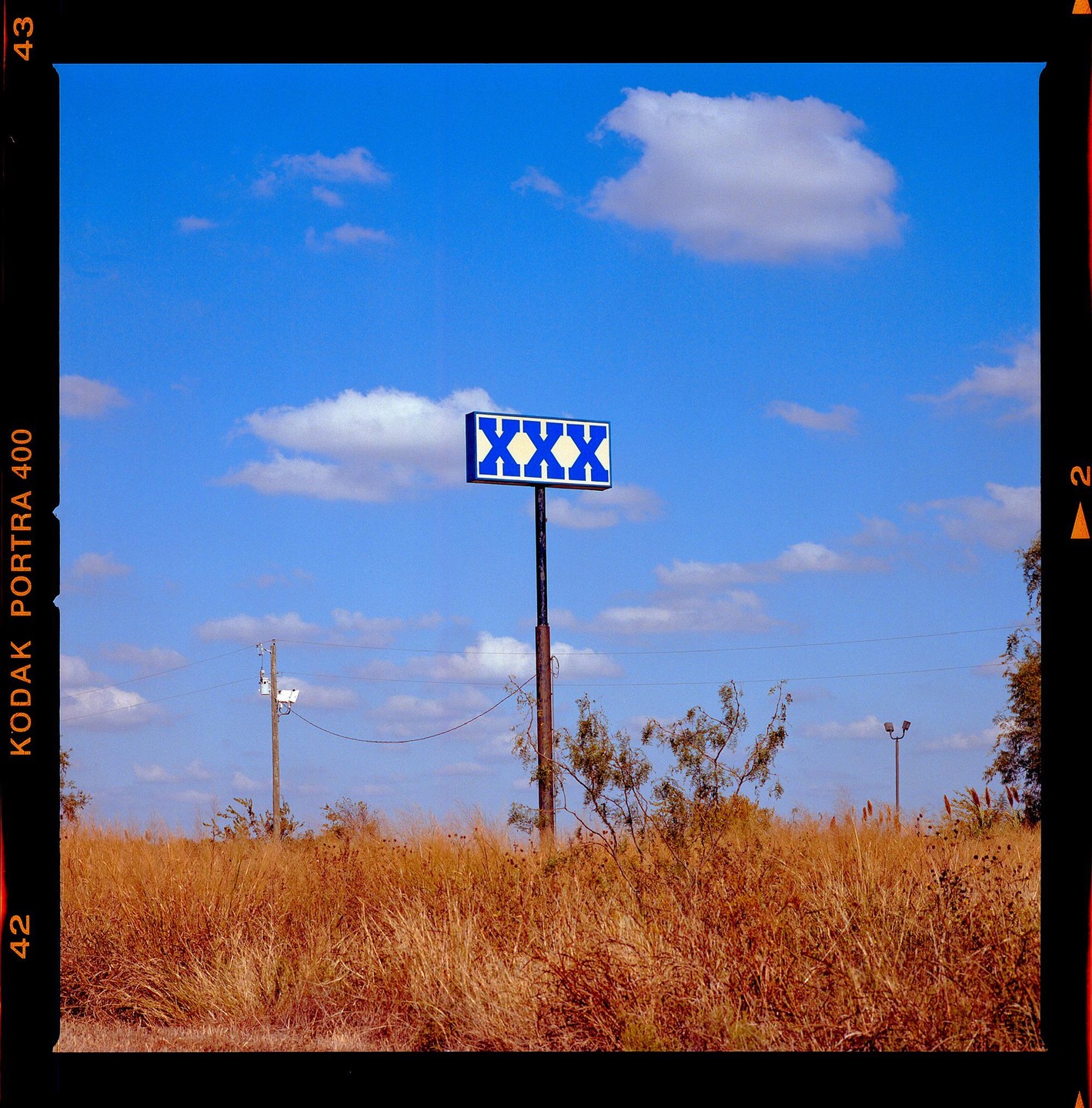 A blue and white XXX sign stands tall above a field of dry grass under a bright blue sky with scattered clouds.
