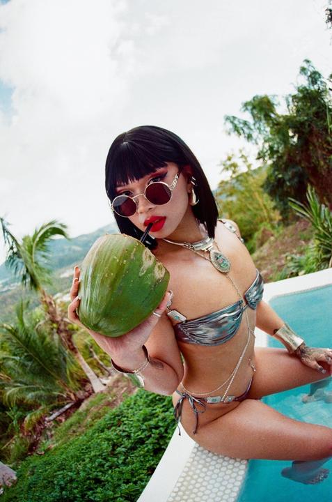 A woman in a bikini and sunglasses sits in a pool, holding a coconut with a straw.