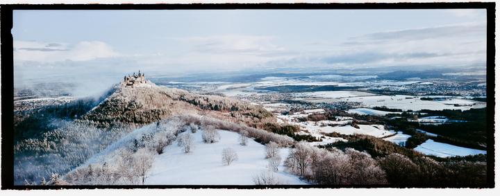 A castle sits atop a snow-covered hill overlooking a vast winter landscape with a town in the distance.