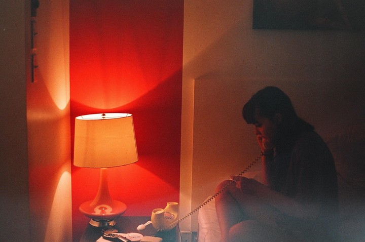 A woman sits on a bed in a dimly lit room, talking on a rotary phone next to a lamp.