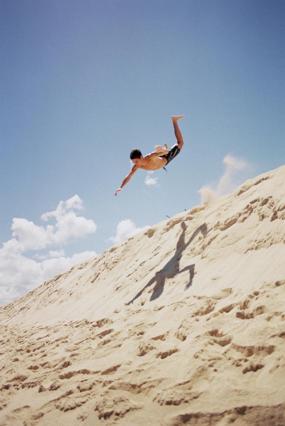 A man jumps in the air above a sand dune, casting a shadow on the sand.