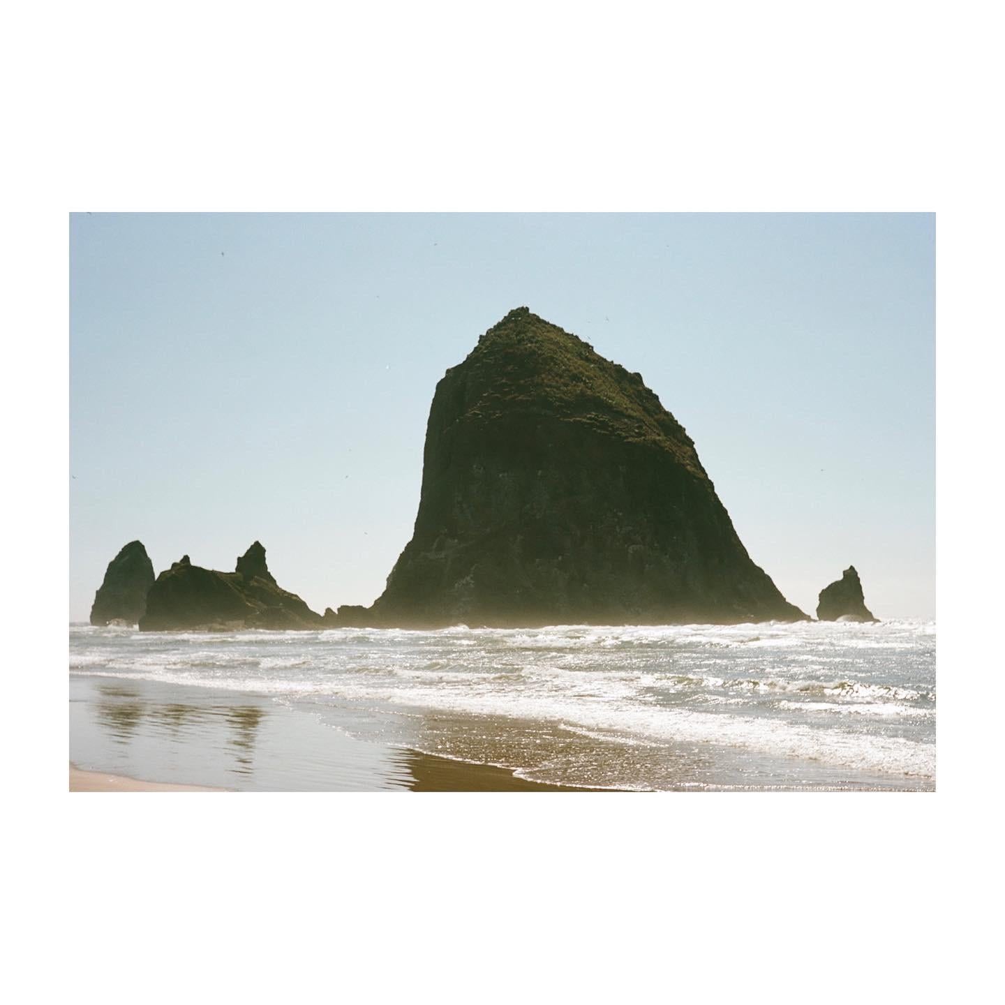 A large rock formation rises from the ocean with waves breaking on the shore.