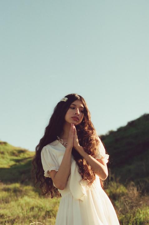 A woman with long, curly hair and a white dress stands in a grassy field with her hands clasped together.