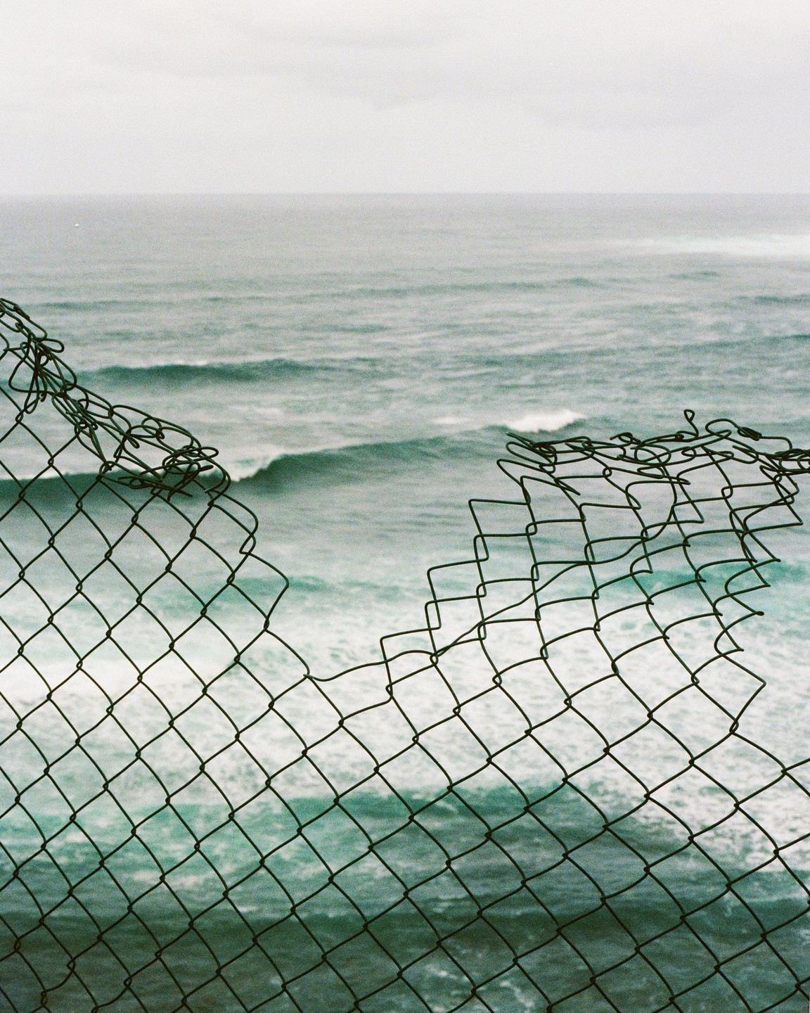 A torn chain-link fence is in the foreground with the Atlantic Ocean and waves in the background.