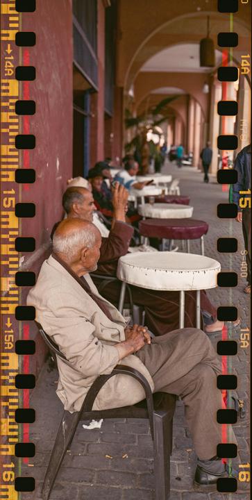 Several men are sitting at small tables along a red-walled arched walkway.
