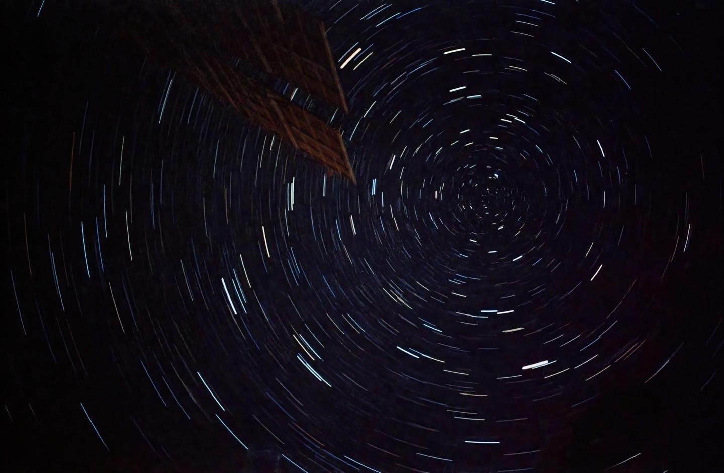 A long exposure photograph captures the circular star trails in the night sky, with a dark structure visible in the upper left corner.