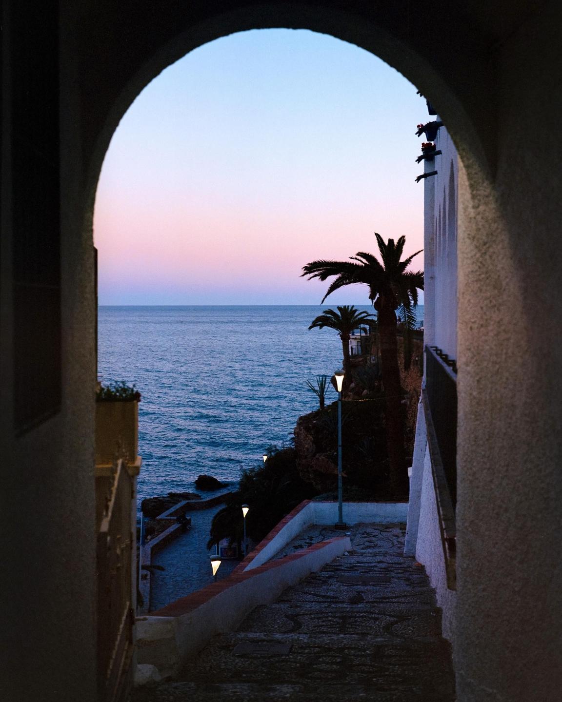 A stone archway frames a view of the ocean and palm trees at sunset.