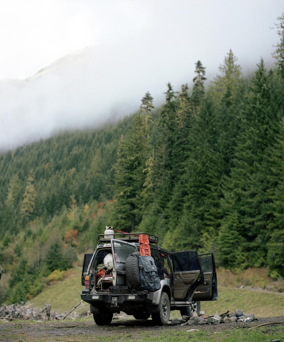 A vehicle with its doors open and camping gear loaded on the roof and in the back is parked on a dirt road in a forest with a hazy mountain in the background.