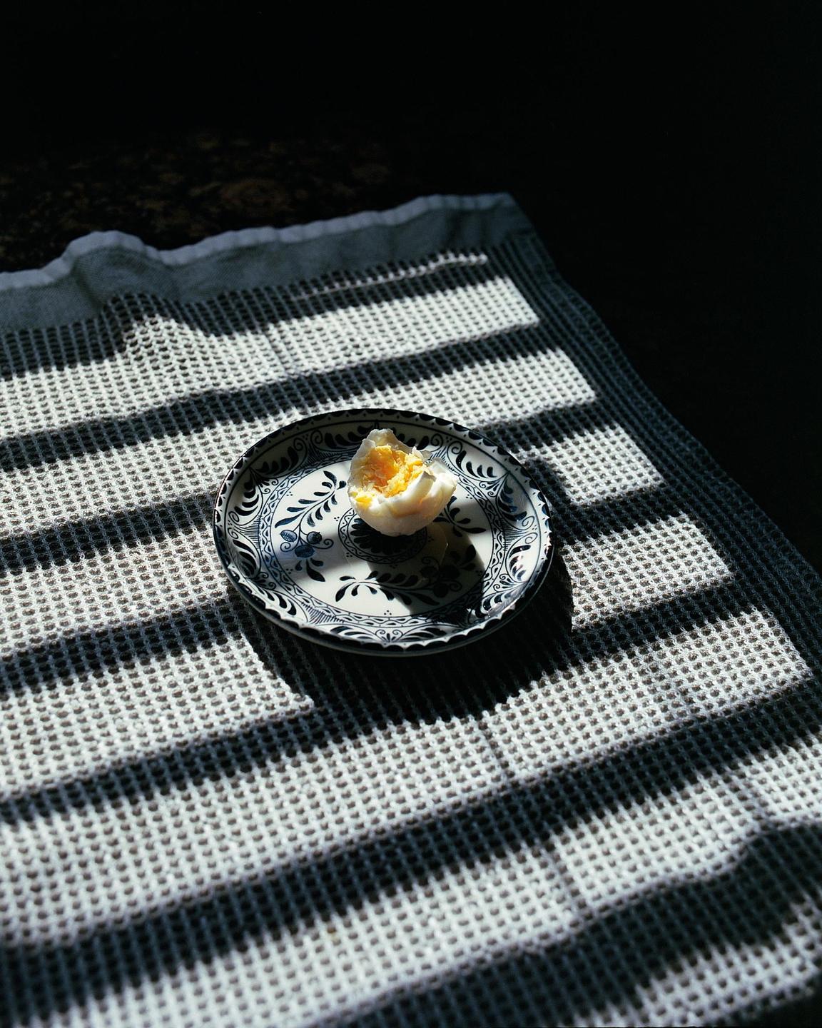 A half-eaten boiled egg sits on a decorative plate on a textured cloth, with strong shadows cast across the surface.