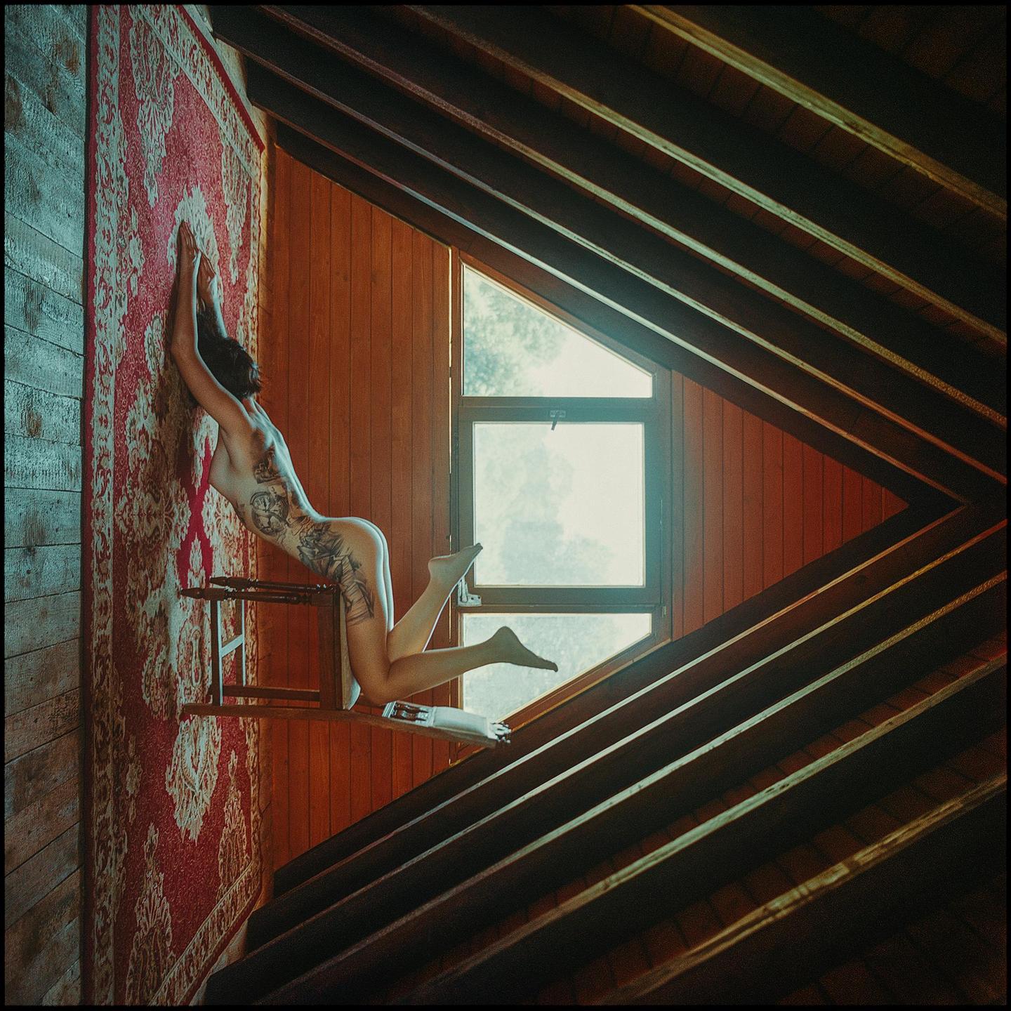 A woman with tattoos on her back and leg is posing on a chair against a patterned rug in a room with wooden walls and a large window.