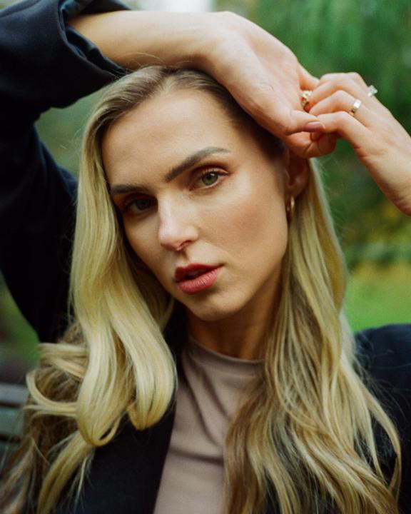 A woman with long blonde hair and red lipstick poses with her hands above her head.