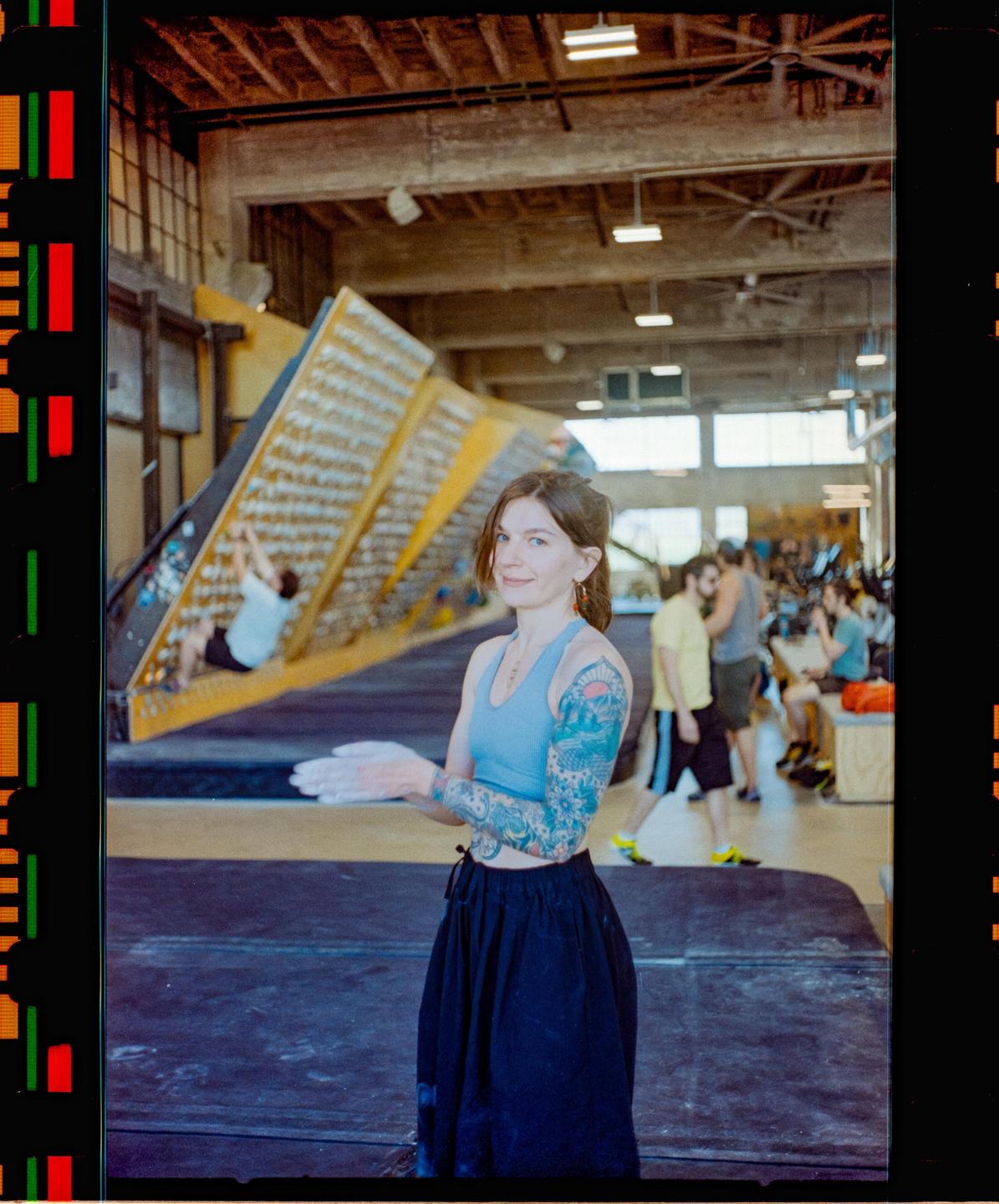 A woman with tattooed arms stands in a climbing gym with chalk on her hands.