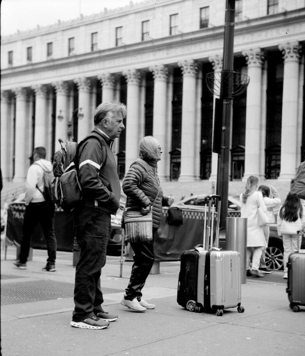A man and a woman stand on a city sidewalk with luggage and a large building with columns in the background.