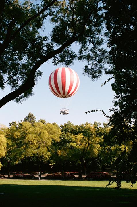 A red and white striped hot air balloon floats above a park with green trees and grass.