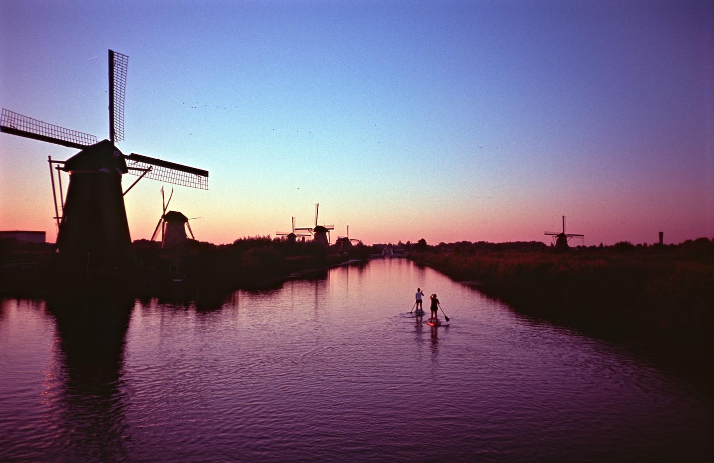 Two people paddleboard on a canal lined with windmills at dusk.
