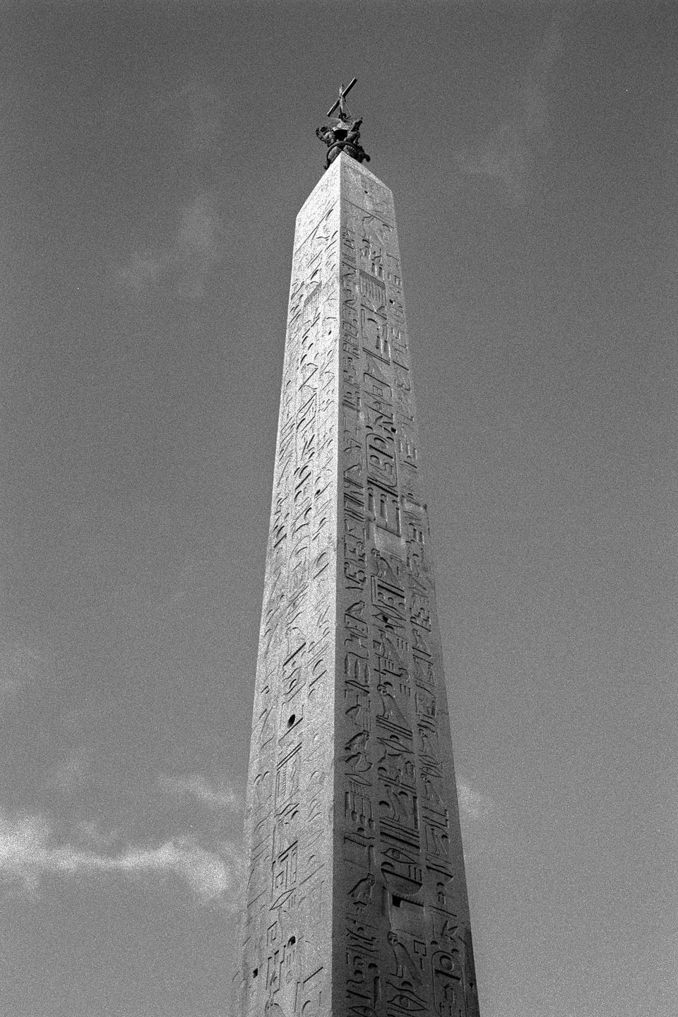 A tall, ancient obelisk with hieroglyphs and a statue on top stands against a bright sky.
