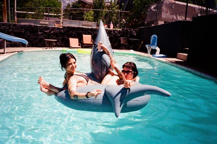 Two women are floating on a shark-shaped inflatable in a swimming pool.