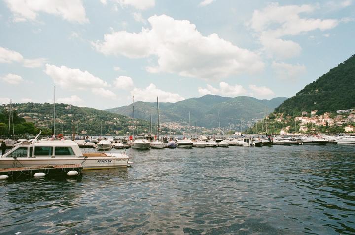 A marina filled with boats is nestled at the base of a lush, green mountain with houses dotting its slopes under a cloudy sky.