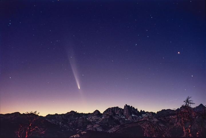 A comet with a bright tail is visible in the night sky above a mountain range.