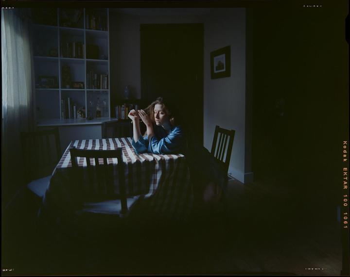 A woman sits at a table with a checkered tablecloth, her hands in her hair, in a dimly lit room with a bookshelf.