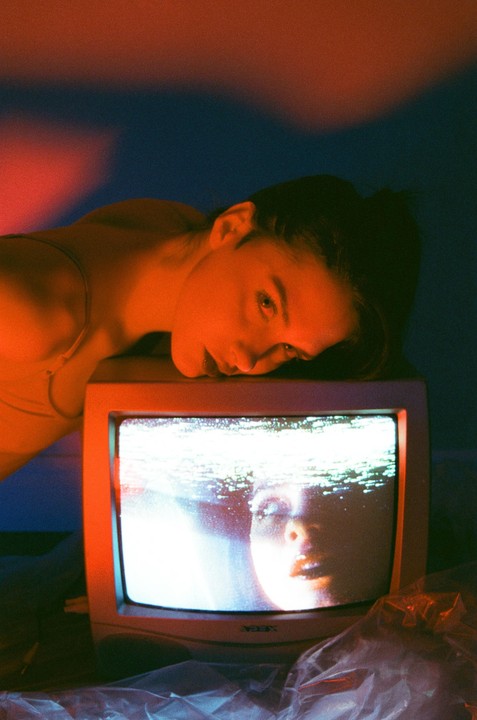 A woman rests her head on a television screen displaying a distorted image of a face.