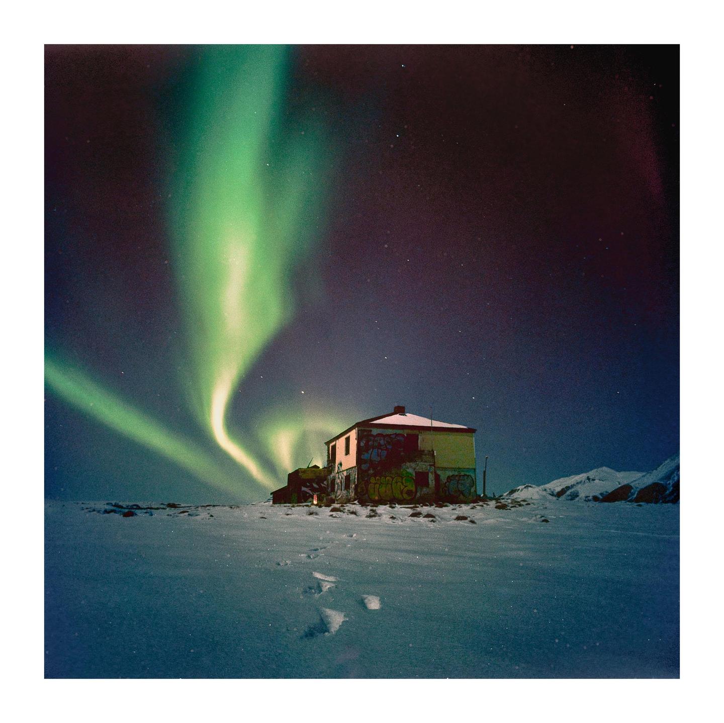 The northern lights glow above a house in a snowy landscape with mountains in the distance.